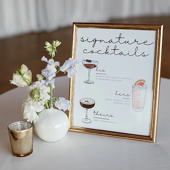 Signature cocktails sign in a gold frame with illustrated his, hers, and theirs drinks beside pastel flowers and a votive on draped backdrop