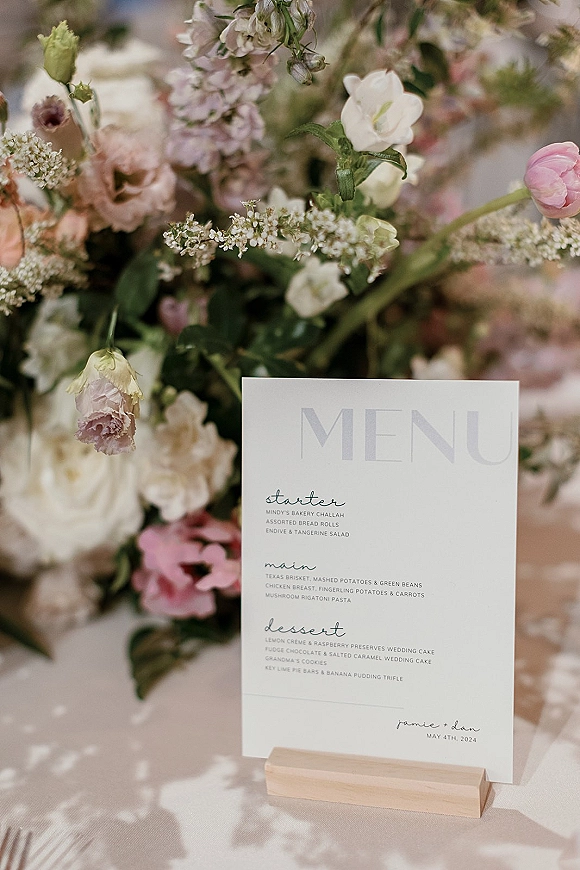 Wedding menu card displayed in a wooden menu holder with greenery accent, styled on a reception table with blurred florals behind
