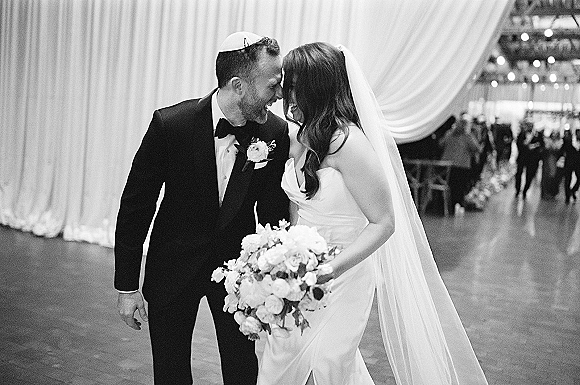 Wedding couple portrait in a black and white wedding photo, bride in strapless dress holding bouquet as groom in kippah touches foreheads indoors with string lights