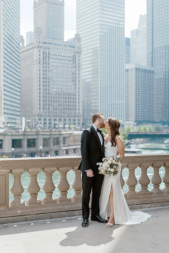 Wedding kiss portrait of bride and groom kissing, she holds a bouquet in a strapless slit gown by a sunlit riverfront skyline and bridge
