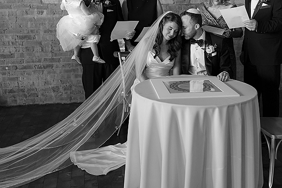 Wedding ketubah signing as bride in a veil and groom in a tuxedo sign papers at a round table, guests by a brick wall indoors