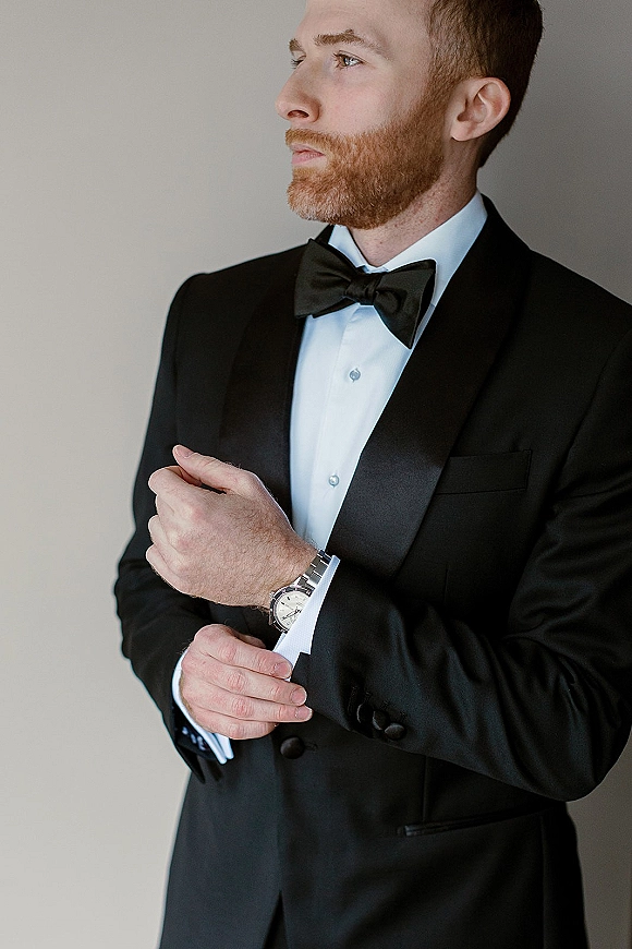 Groom portrait in a black tuxedo, bow tie, and white dress shirt, showing cufflinks and wristwatch against a neutral wall