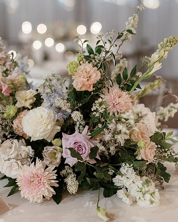 Wedding centerpiece flowers in a pastel wedding centerpiece of roses, chrysanthemums, and greenery on a reception table with soft bokeh lights