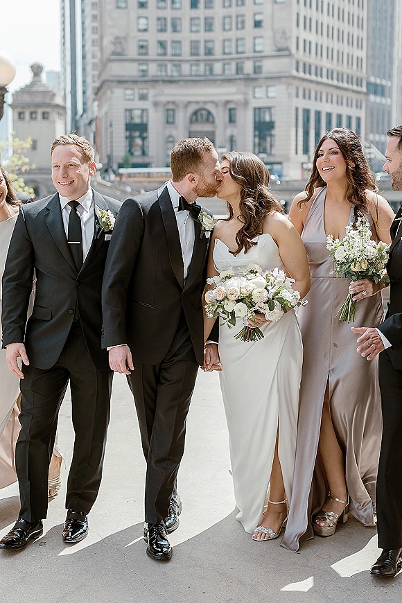 Wedding party portrait of bride and groom kissing as the bridal party walks on a city street between tall buildings in daylight