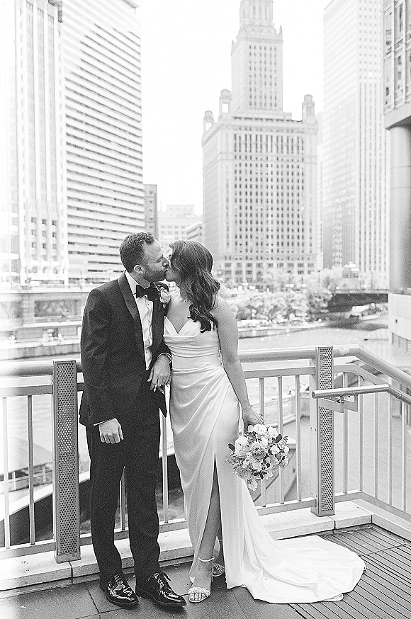Wedding kiss portrait of bride and groom kissing on a rooftop terrace railing, her strapless satin gown and bouquet set against the city skyline
