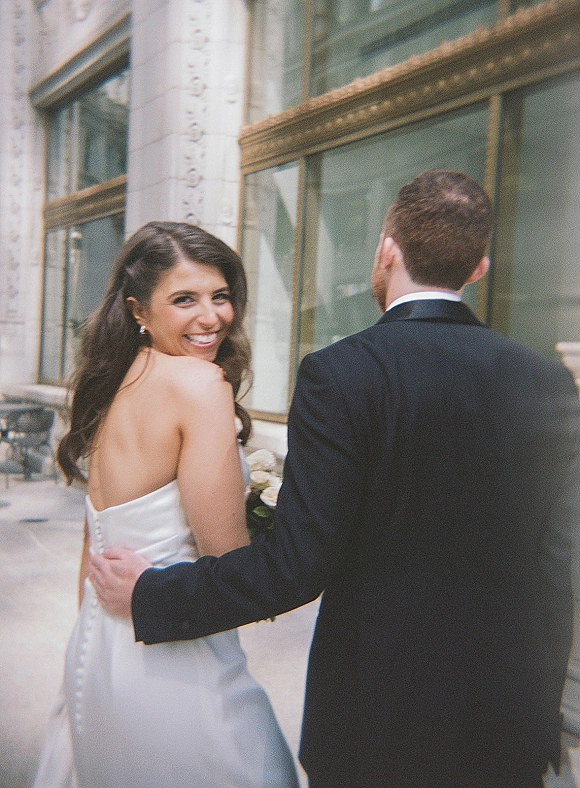 Couple portrait of bride looking back while walking with groom arm around her, holding a white rose bouquet beside city building windows
