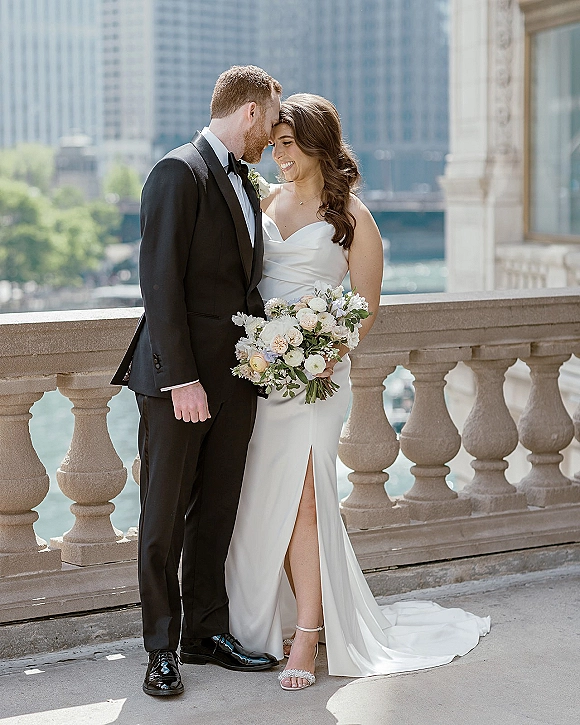 Couple portrait of bride in a strapless slit dress holding a bouquet, leaning into groom in black tuxedo on a stone balcony above a river