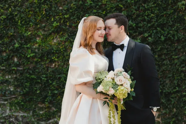 Couple portrait of bride and groom holding a bouquet, her long veil draped over a satin dress beside an ivy wall greenery backdrop