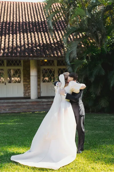Wedding hug as bride and groom embrace on a grass lawn, her cathedral veil and train flowing, palm trees and tiled-roof villa behind