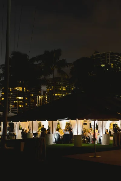 Wedding reception tent with string lights glowing over round tables and white draping, set on a lawn with palm trees and city buildings at night