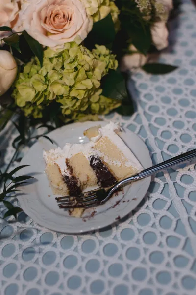 Wedding cake slice with white frosting and chocolate filling on a cake cutting dessert plate and fork beside roses centerpiece on patterned tablecloth