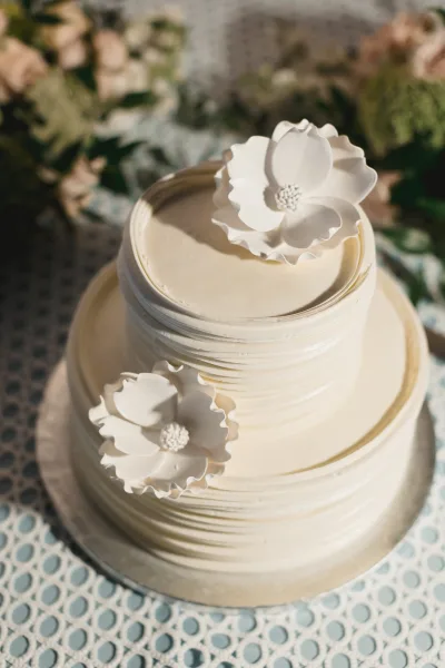 Wedding cake with buttercream frosting on a patterned tablecloth, topped with white fondant flowers against blurred greenery and florals