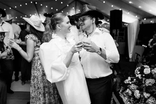 Wedding reception moment as bride and groom laugh and toast drinks, groom in cowboy hat under string lights in a crowded tent
