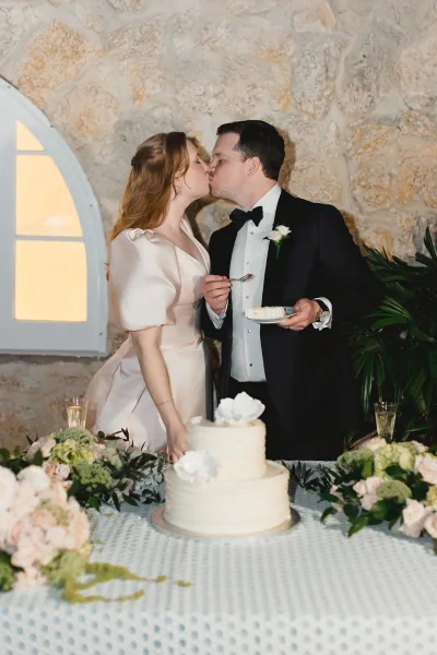 Cake cutting moment with bride and groom cake cutting at a two-tier white buttercream cake table, stone wall and arched window behind