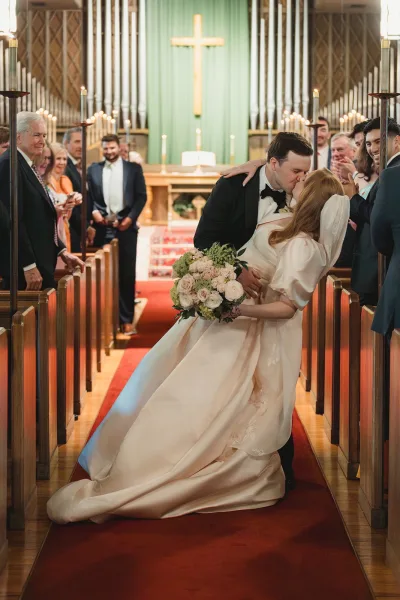 Wedding kiss portrait of bride and groom kissing in the aisle with bouquet and veil, framed by tall candles, red runner, and church altar cross