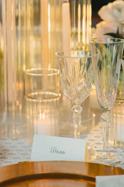 Reception tablescape with wedding place card, crystal wine glasses, gold charger plate, taper candles and florals on a textured cloth, bokeh lights behind