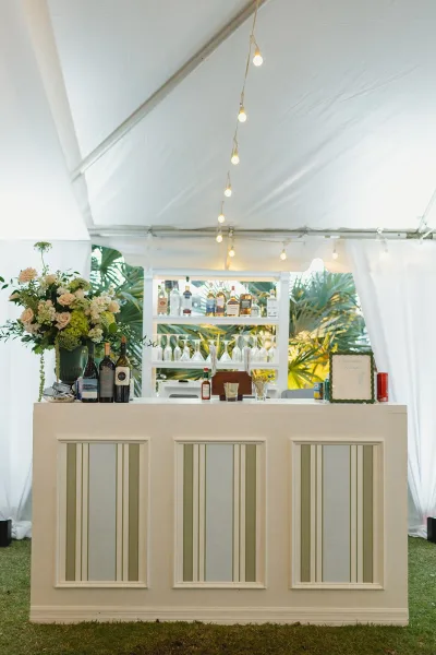 Wedding bar setup with a white bar counter lined with liquor and wine bottles, glassware, and floral accents under tent string lights