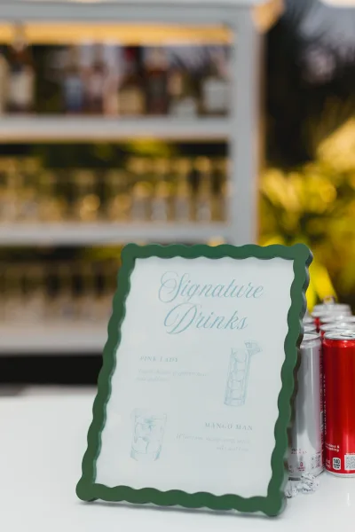 Signature drinks sign in a scalloped frame with illustrated cocktails and soda cans, displayed on a bar shelf with bottles and greenery in outdoor light