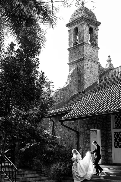 Wedding couple portrait of bride and groom on steps, she holds bouquet as he lifts her dress train, with veil by a stone chapel bell tower