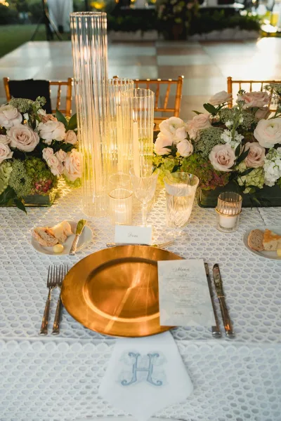 Reception tablescape with wedding table centerpiece of tall cylinder vases and floating candles, blush roses, and gold charger plate in draped tent by checkered dance floor