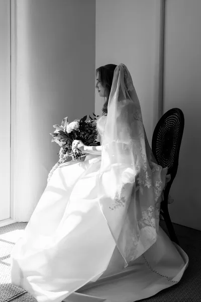 Bridal portrait of a bride sitting by window, holding a white flower bouquet with lace veil details and strapless gown in soft light