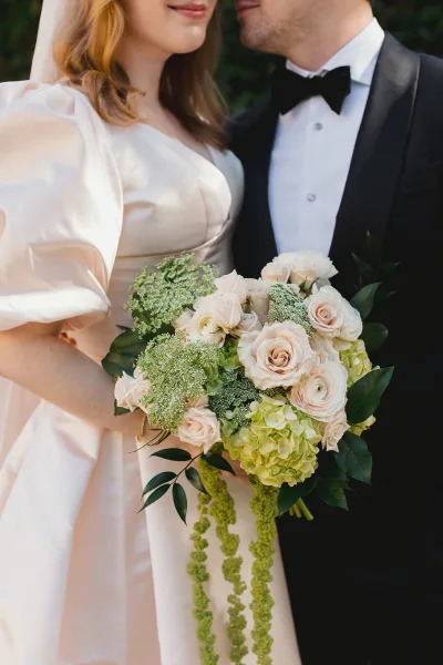 Couple portrait of bride and groom close up, holding a blush rose bouquet, her veil and satin puff-sleeve dress against greenery backdrop