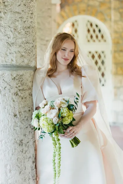 Bridal portrait of a bride holding bouquet with a cathedral veil, off-shoulder dress and cascading roses by a stone archway column