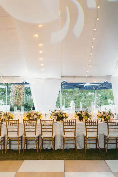 Reception tablescape with long head table decor on white lace tablecloth, white roses and greenery, candles in tall glass vases under string lights in a draped tent