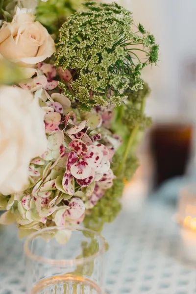 Wedding centerpiece of white roses and hydrangea with lush green florals beside a glass votive candle on a table, bokeh reception lights behind