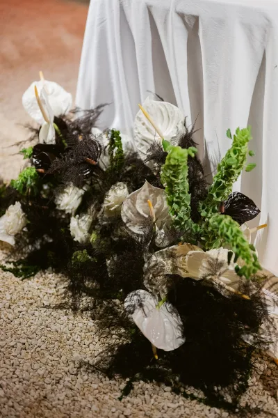 Ceremony florals in a ground floral arrangement with white anthurium and roses, black foliage, and white draped fabric on gravel aisle