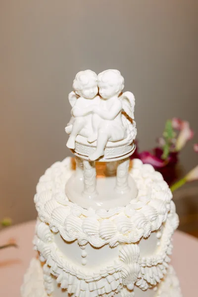 Wedding cake with vintage wedding cake styling, white-on-white ruffle piped frosting and cherub figurine topper against a neutral wall with blurred flowers