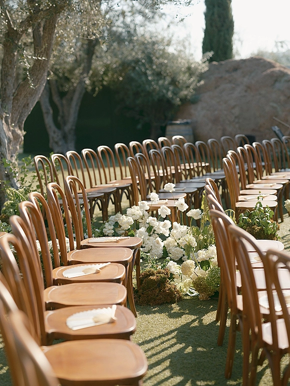 Ceremony aisle decor with low white flowers and greenery lining a grassy lawn aisle between wood chairs, folded fans in sunlight