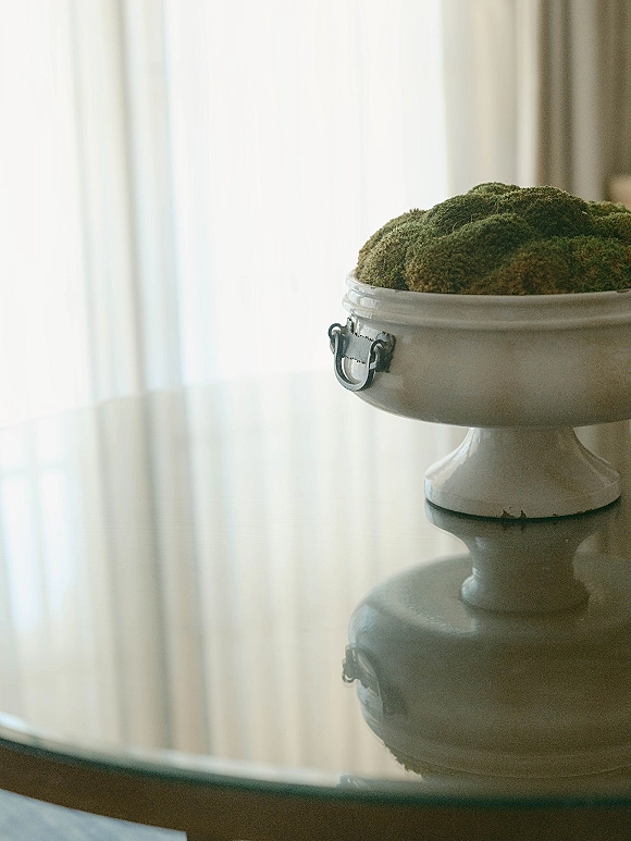 Moss centerpiece in a white pedestal bowl with metal handles on a glass tabletop, softly lit by window light and sheer curtains