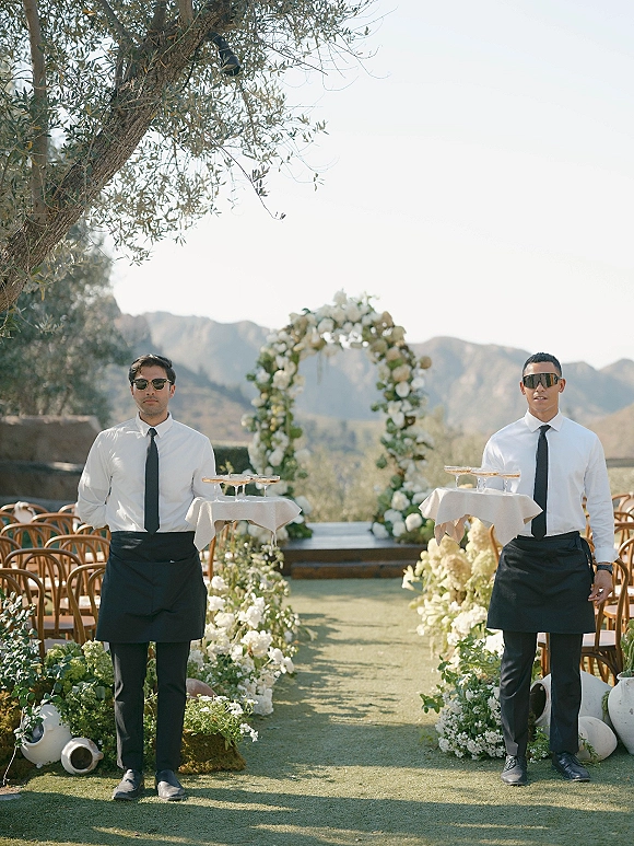 Ceremony setup for an outdoor wedding ceremony with a floral arch, white blooms and greenery, wooden chairs on a mountain-view terrace.