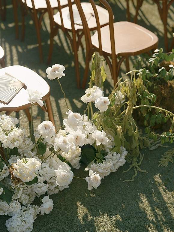 Ceremony aisle flowers in white roses and hydrangeas with greenery along a sunlit grass lawn beside wooden chairs with fans