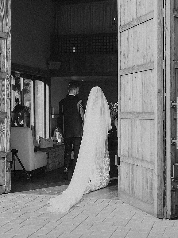 Wedding processional as bride walking down aisle beside groom, her long veil and dress train trailing on brick pavers through wooden doors