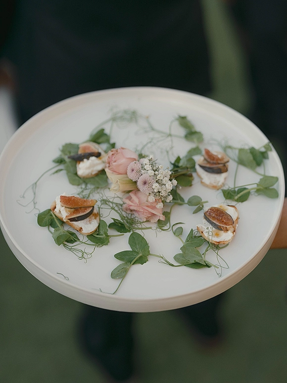 Wedding appetizers on a white serving platter with fig and cheese bite-size crostini, edible greens, and pink rose garnish, guests blurred behind