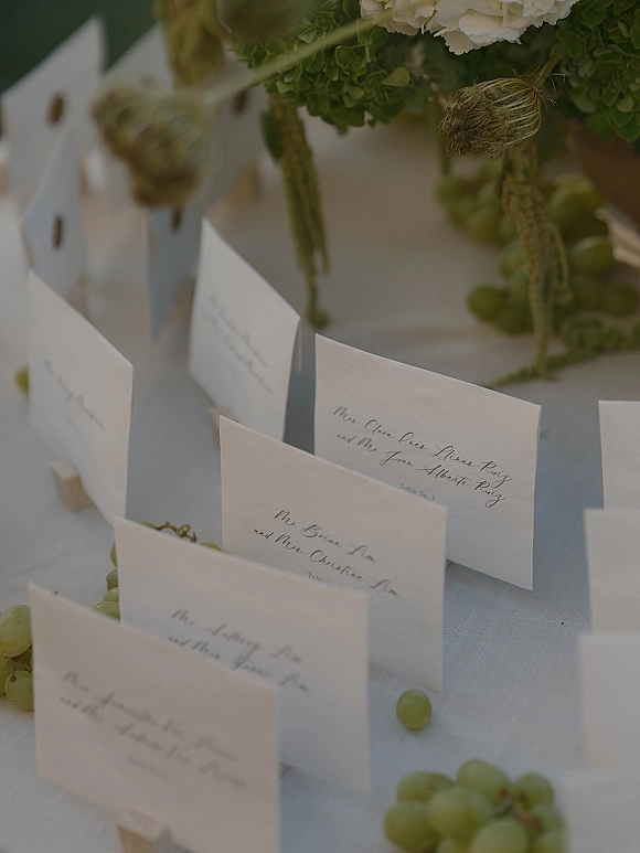Wedding place cards with calligraphy place cards in wooden stands beside grapes and a green-and-white floral centerpiece on a linen tablecloth