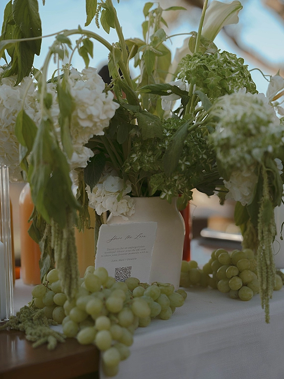 Reception tablescape with wedding floral centerpiece of white hydrangeas in a ceramic vase, grapes and candle glass, with blurred outdoor guests