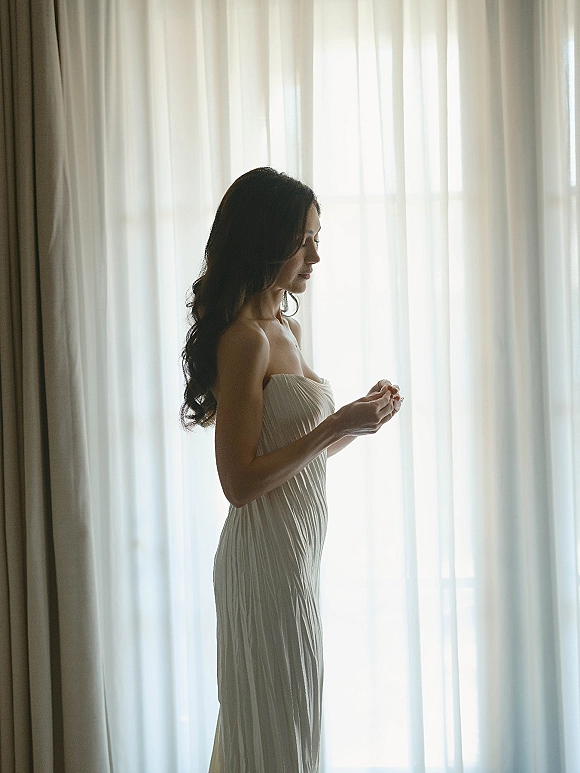 Bridal portrait of a bride getting ready by window light, adjusting her ring in a strapless pleated gown beside sheer curtains