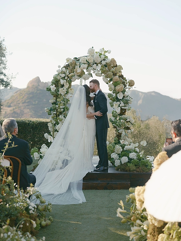 Ceremony kiss as bride and groom embrace beneath a white floral arch, long veil flowing on a platform with mountains behind guests
