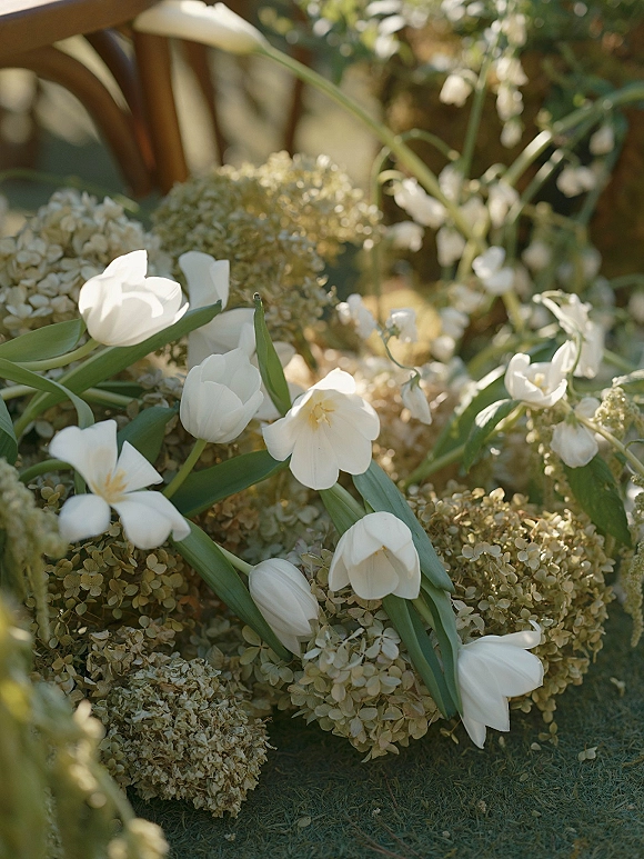 Wedding florals of white tulip wedding flowers and hydrangea with greenery on the ground beside a wooden chair in garden grass