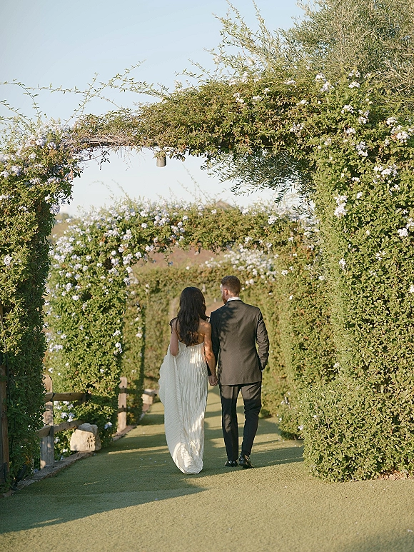 Couple portrait of bride and groom walking away holding hands beneath a white flower archway on a garden pathway under blue sky