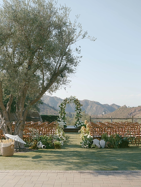 Ceremony setup for an outdoor wedding ceremony with a white flower and greenery arch, aisle florals, wooden chairs, and mountain views behind