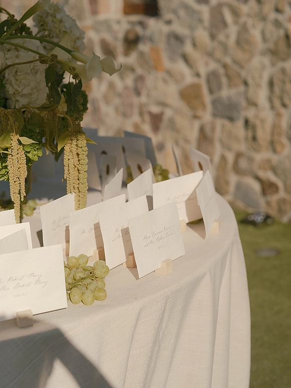 Wedding place cards in wooden holders on a place card table with white linen, hydrangea and hanging amaranthus, greenery and grapes by a stone wall