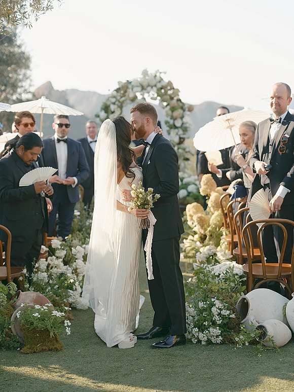 Wedding kiss as bride in strapless gown holds bouquet, long veil flowing down garden aisle under floral arch with mountains beyond