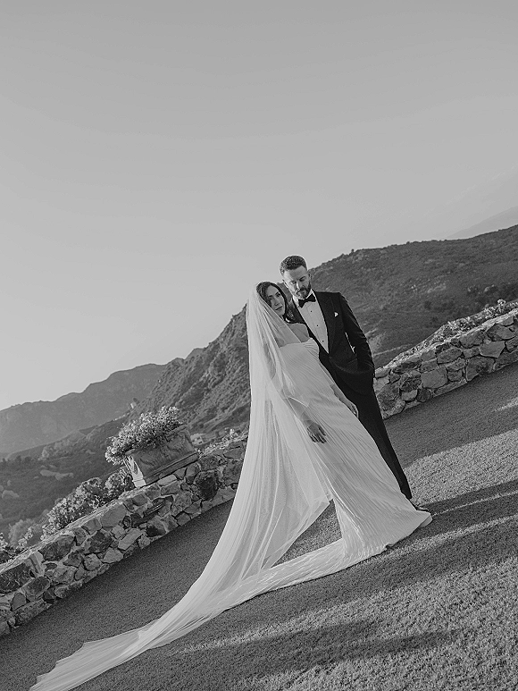 Couple portrait of bride in strapless gown with long wedding veil train leaning on groom in tuxedo by a stone wall with mountains beyond