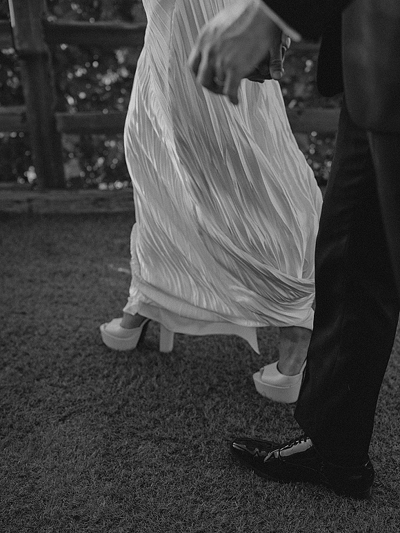 Wedding couple detail of bride and groom holding hands as they walk on a grassy lawn, showing dress hem, white platform heels, and suit shoes