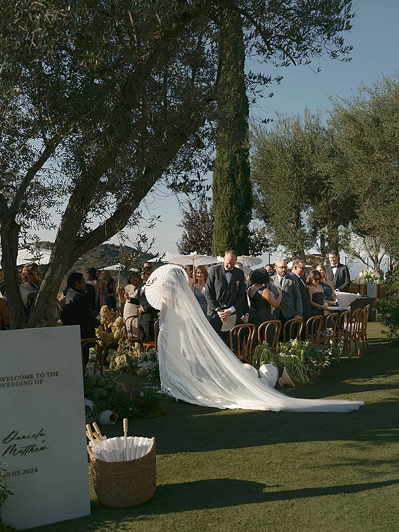 Wedding processional with bride walking down aisle in a long veil and train, guests under white parasols on an outdoor lawn with olive trees