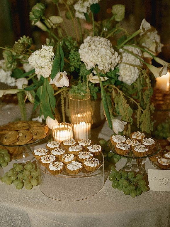 Wedding dessert table with mini tarts and cookies on glass stands, a white floral centerpiece, and candlelit ribbed votives in dim reception light